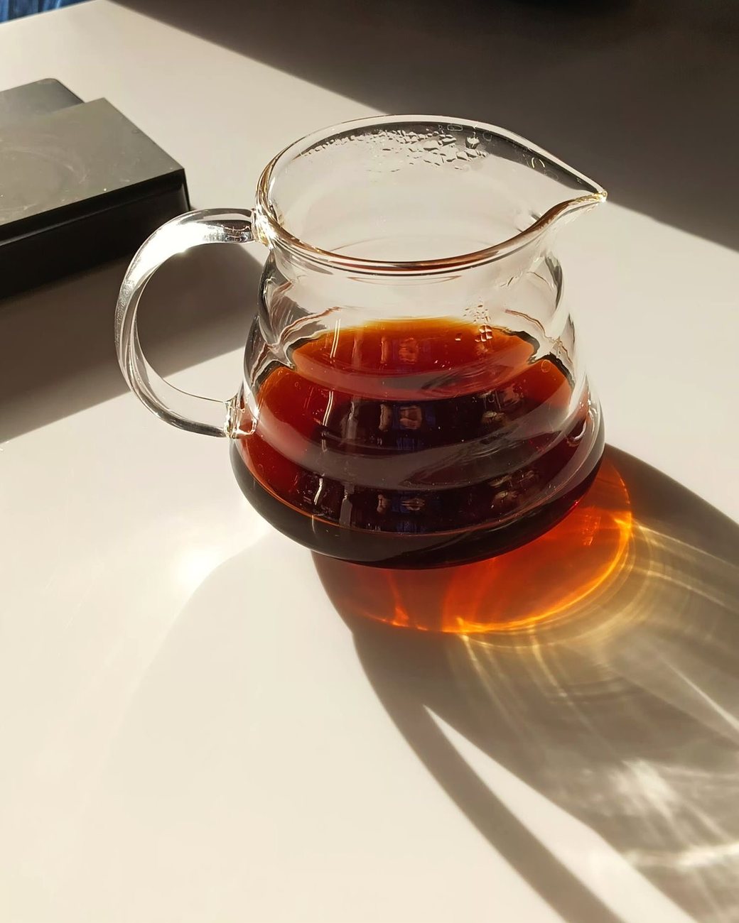 Jarra de vidro com café coado sob a luz do sol, projetando reflexo âmbar na mesa